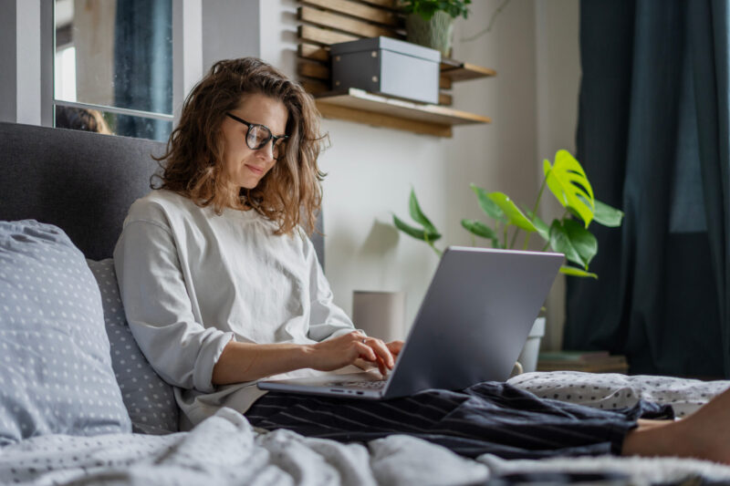 Young beautiful smiling woman working on business project using laptop while sitting in bed at home Junge Frau, die mit Laptop an einem Geschäftsprojekt arbeitet, während sie im Bett liegt